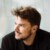 Portrait of Danish star architect Bjarke Ingels, looking left, wearing a black shirt, beard, in front of a grey, glassy background.