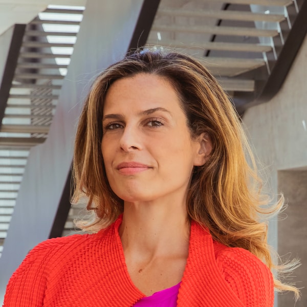 Portrait image of economist Francesca Bria standing outdoors in front of a metal staircase, wearing a red jacket over a purple shirt, looking at the camera.