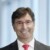 Headshot portrait of Niko Mohr, member of the Executive Board at Friedhelm Loh Group, standing in front of a grey-blue background, smiling at the camera, wearing red-rimmed glasses.