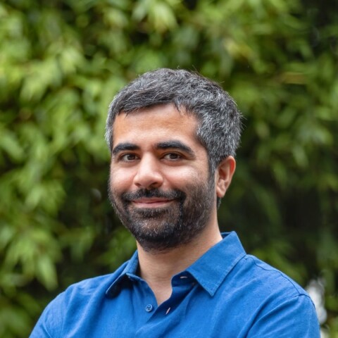 Outdoor portrait of web3 entrepreneur Herman Narula, co-founder and CEO of Improbable, wearing a blue shirt, standing in front of a lush green background in nature.