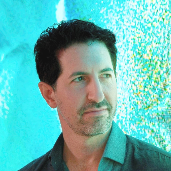 Colorful portrait of neuroscientist Moran Cerf, seen in front of a blue-green digital screen, looking over his left sholder, outside the frame.