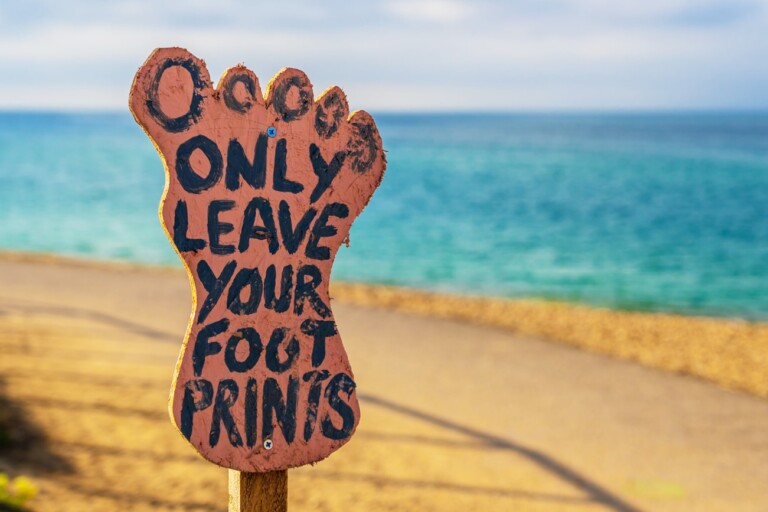 Photo illustrating sustainability, shows a beach and a wooden sign, shaped like a foot, that says, “Only leave your footprints”