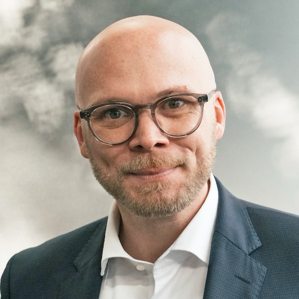 Headshot portrait of Fabian Mehring, State Minister Bavarian Ministry for Digital Affairs, in front of a blurry, grey backgound, smiling at the camera, wearing a suit and glasses.