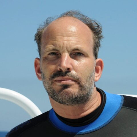 Portrait image of nature photographer Hussain Aga Khan, wearing a diver’s suit, presumably on a boat out on the ocean.