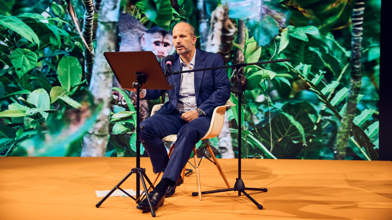 Nature photographer Prince Hussain Aga Khan speaks about the beauty of nature at the DLD conference in Munich. He sits at a pedestal, with a display behind him that shows a rainforest and monkey.