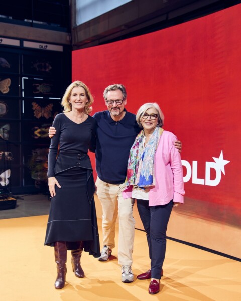 Group photo of DLD Nature hosts Maria Furtwängler, Eckart v. Hirschhausen and Steffi Czerny on stage, in front of a red poster showing the DLD logo (white)