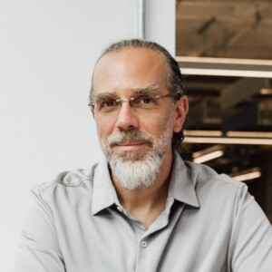 Headshot portrait image of technology pioneer Astro Teller, director of Google X, seen in an office biulding in front of a grey wall, smiling at the camera.