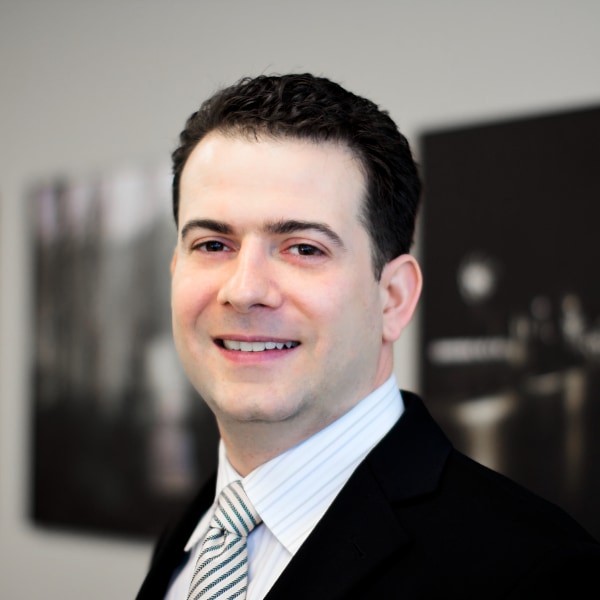 Portrait image of quantum computing specialist Michele Mosca, CEO & President of evolutionQ Inc., wearing a suit, standing in front of a wall with black-and-white photos blurry in the background.