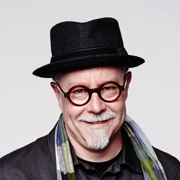 Portrait image of IDEO chairman Tim Brown, wearing a black hat, glasses and a colorful scarf, smiling at the camera.