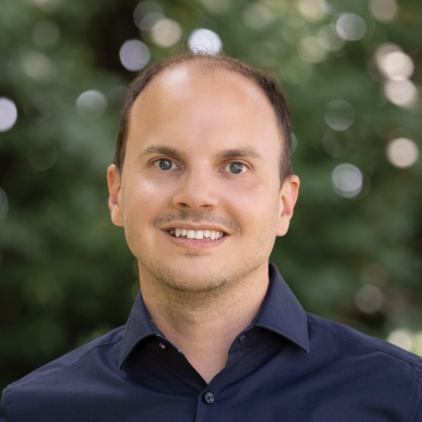 Outdoor portrait of Dr. Alexander Glätzle, CEO and co-founder of planqc, standing in front of green trees, visible blurry in the background.