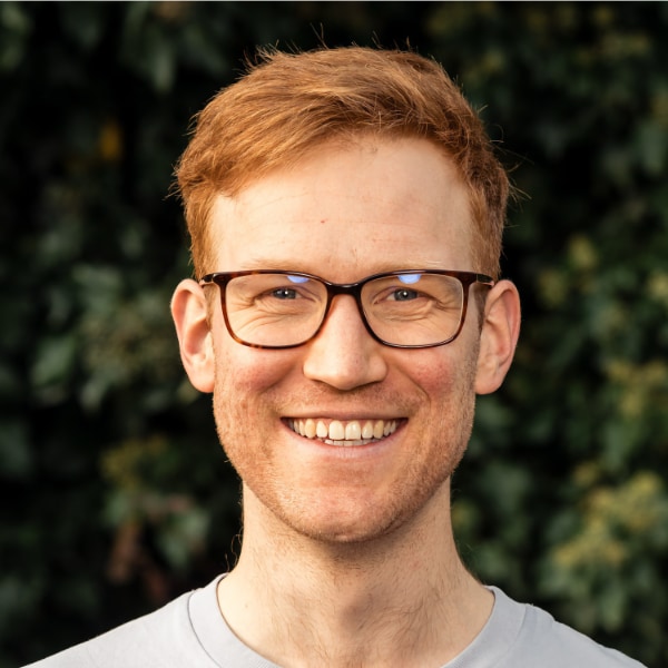 Environmental portrait of Black Forest Labs co-founder Andreas Blattmann, wearing glasses and a grey t-shirt, seen outdoors in front of a natural green background.