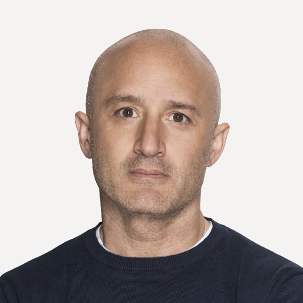 Close-up portrait image of artist and photographer Andrew Zuckerman, wearing a black pullover, standing in front of a white background.