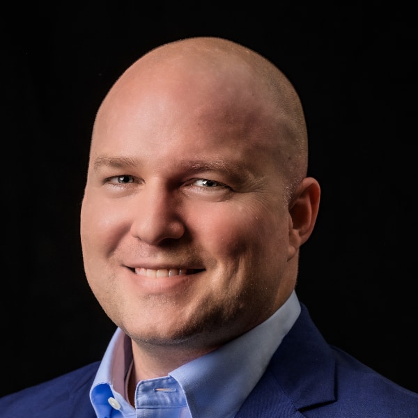 Studio portrait of climate-tech excutive Bryan Hassin, CEO of DexMat, smiling at the camera, head brightly lit in front of a black background.