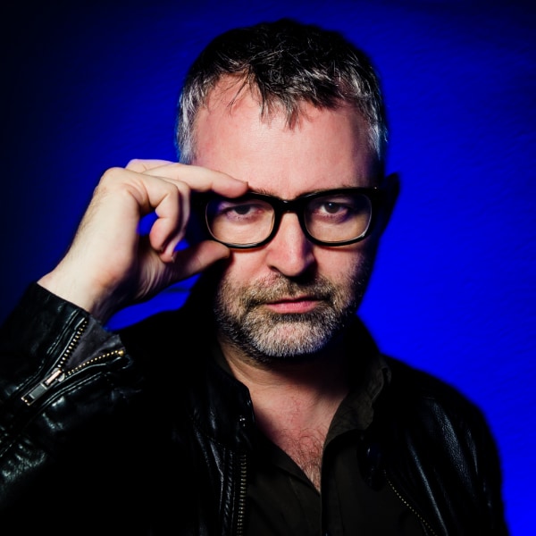 Colorful portrait image of tech journalist Mike Butcher, wearing a black leather jacket, putting his right hand on his glasses, standing in front of a purple studio background.