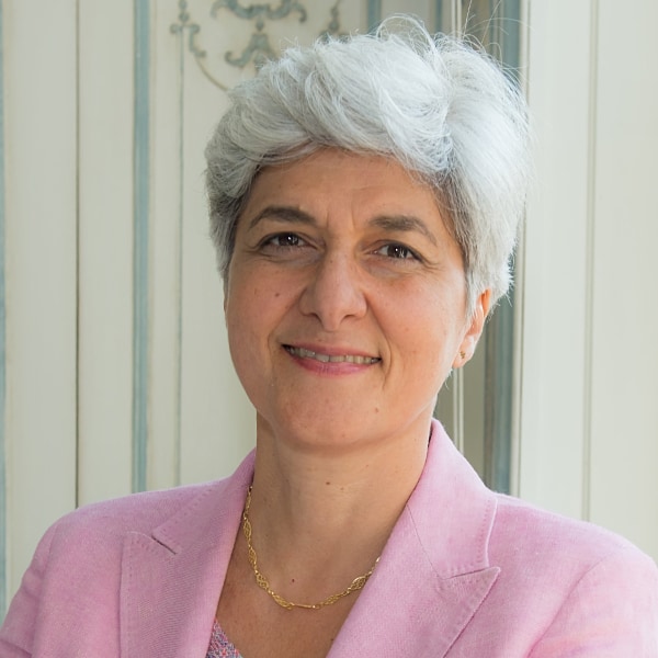 Indoor headshot portrait of Sylvie Goulard, smiling at the camera, wearing a pink jacket, standing in front of a grey wall with green and blue stripes on the wallpaper.