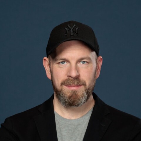 Studio portrait of Serviceplan manager Alex Turtschan, wearing a black baseball cap, looking at the camera, standing in front of a dark blue background.