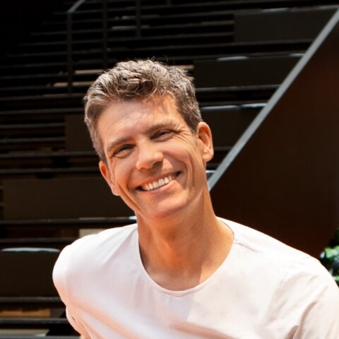 Environmental portrait of Alexander Nagel, Managing Partner and Creative Lead, Serviceplan Make, sitting in front of a set of stairs, wearing a white t-shirt, smiling at the camera.