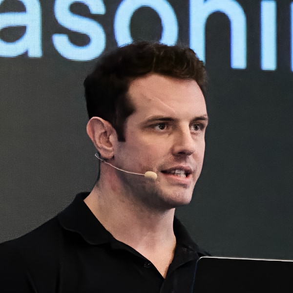 Photo of OpenAI manager Colin Jarvis, wearing a microphone headset, speaking at a conference with blue letters visible in the background.