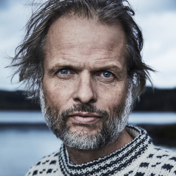 Dramatic headshot portrait of Norwegian explorer Erling Kagge, seen outdoors in front of a cloudy, blue-grey sky, looking at the camera, hair tousled.