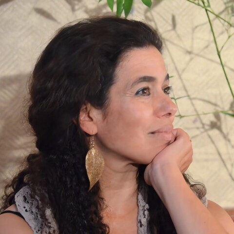 Biology professor Farhah Assaad sitting in front of a wall, looking to her left, outside the frame, smiling.