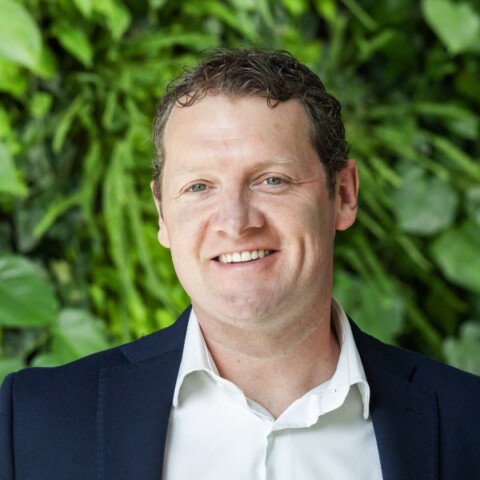 Outdoor portrait of Ryan Archer, co-founder of indoor-farming company Horizon Growers, seen in front of a plant with green leaves, smiling at the camera.