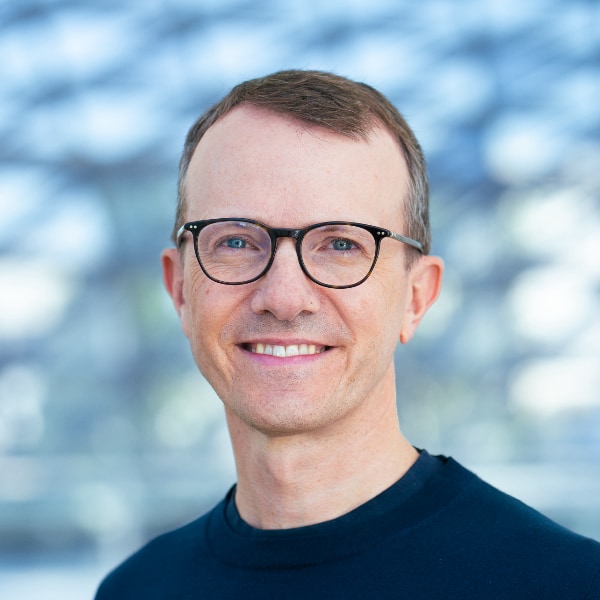 Headshot portrait of Sebastian Dettmers, CEO of recruiting plattform Stepstone, smiling at the camera, seen before a blurry blue pattern background.