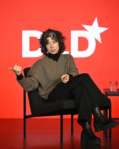 Meredith Whittaker, Signal Foundation, speaks at the DLD Munich 2025 conference. She’s sitting on a black chair, gesticulating, in front of a red background showing the white DLD logo.