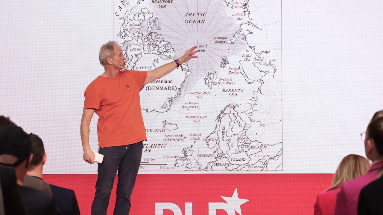 Norwegian explorer Erling Kagge speaks at the DLD Munich Conference 2025, pointing to a map of the Arctic Circle. In the foreground, audience members sit on both sides of the aisle, listening.