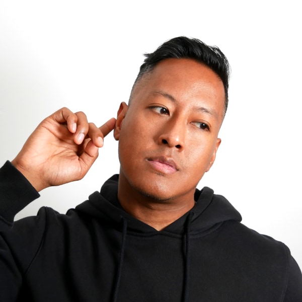Studio portrait of Angelo Tun, aka DJ Angelo, tilting his head to the right, holding one finger of his left hand to his ear, as if listening to a good tune.