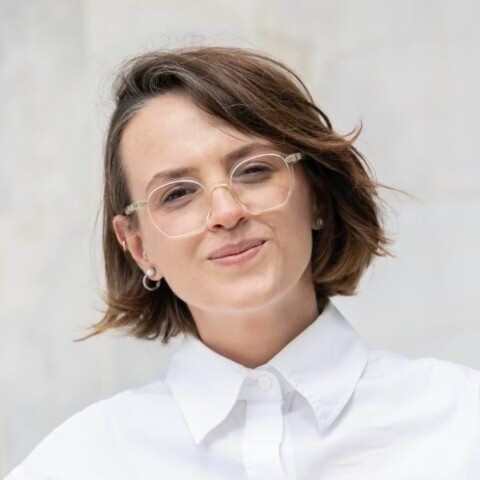 Studio headshot portrait of Helga Mustafaraj, Senior Project Manager Innovation Hub, Tirana, Albania, smiling at the camera in front of a neutral grey background.