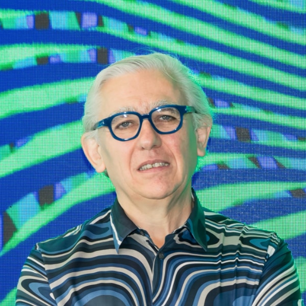 Headshot portrait of artist Miguel Chevalier, wearing a striped blue shirt in front of a digital screen showing a dark blue-cyan pattern.