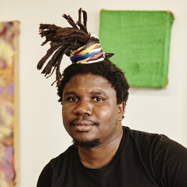 Colorful headshot portrait of artist Alvaro Barrrington, wearing his hear, in dreadlocks, up, smiling at the camera.