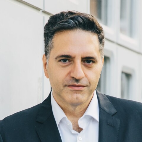 Environmental headshot portrait of Aleksandar Stojanovic, DARI Foundation, standing in front of a grey building wearing a dark suit.