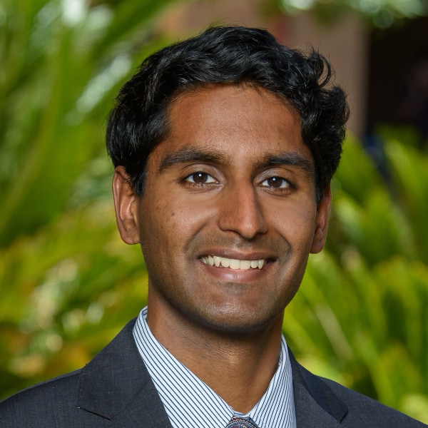Environmental headshot portrait of Stanford University economist Bharat Krishnan Chandar, seen in front of a natural green background, smiling at the camera.
