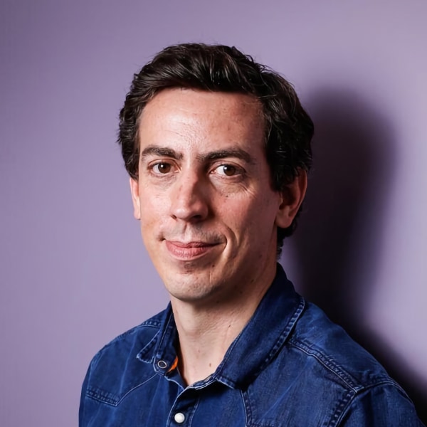 Studio headshot portrait of investigative journalist Laurent Richard, founder of Forbidden Stories, leaning against a purple background, smiling at the camera.