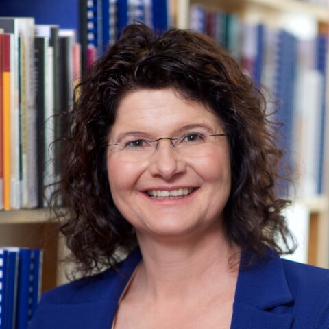 Headshot portrait of journalist Maya Götz, German expert for childrens&rsquo; programming on TV, seen standing in front of a bookshelf, smiling at the camera.