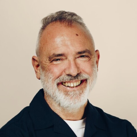 Studio heashot portrait of Nicola Palmarini, also known as Nic Palmarini, in front of a beige background, smiling at the camera.
