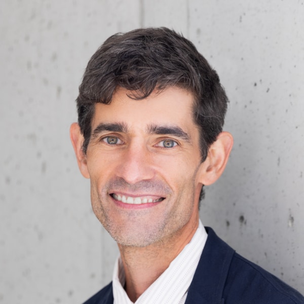 Headshot portrait of journalist Nicholas Thompson, CEO of The Atlantic, standing in front of a grey wall, smiling at the camera.