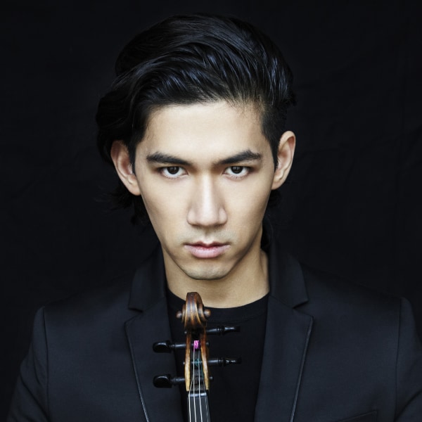Portrait of musician Iskandar Widjaja in front of a black studio background, holding a violin, looking straight at the camera.