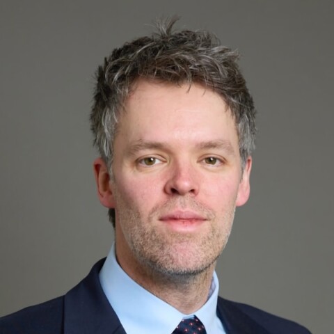 Headshot portrait of British politician Liam Booth-Smith, advisor to Anthropic, in front of a grey studio background.