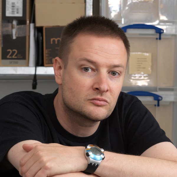 Casual snapshot-style portrait of artist Simon Denny in front of a shelf.