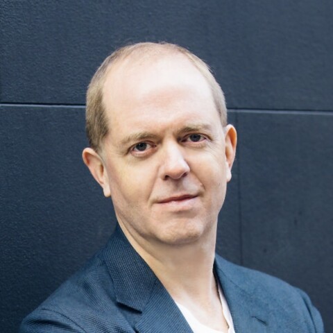 Headshot portrait of UnlikelyAI founder William Tunstall-Pedoe, leaning against a dark blue wall.