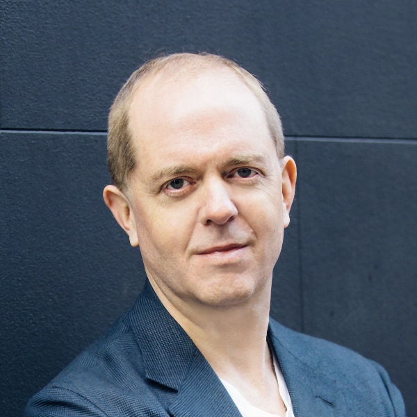 Headshot portrait of UnlikelyAI founder William Tunstall-Pedoe, leaning against a dark blue wall.