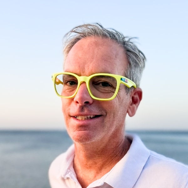 Outdoor portrait of Mike Marqués, Founder & Principal of SHFT Sports, standing by the ocean, wearing bright yellow glasses, smiling.