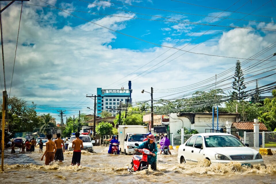 A city in the Global South after a flood. People push bikes through the streets, next to cars driving through high water.